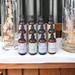 Twelve bottles of high-end lotions by Whole Self Aromatherapy atop a soft white table linen with elegant wedding decorations on either side.